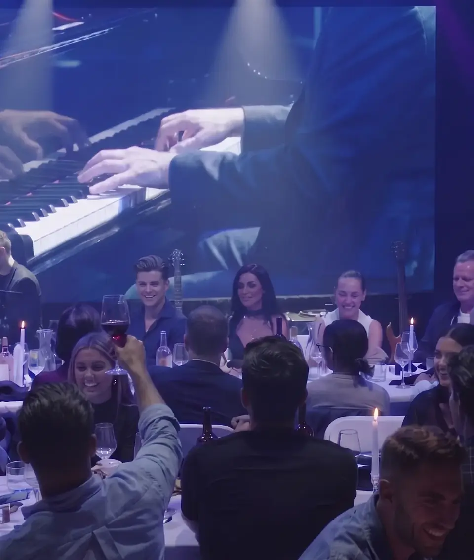 Guests at dinner tables during Piano Wine Night, the pianist's hands on the screen above